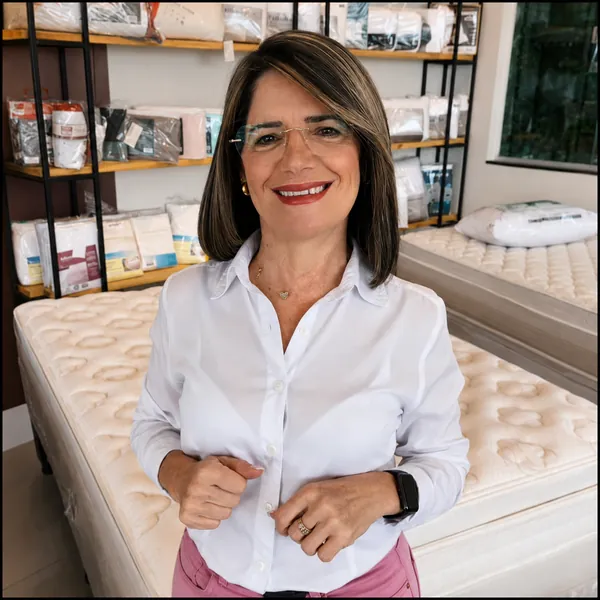 Atendente da Casa dos Colchões sorrindo entre a prateleira de enxoval e uma cama box montada